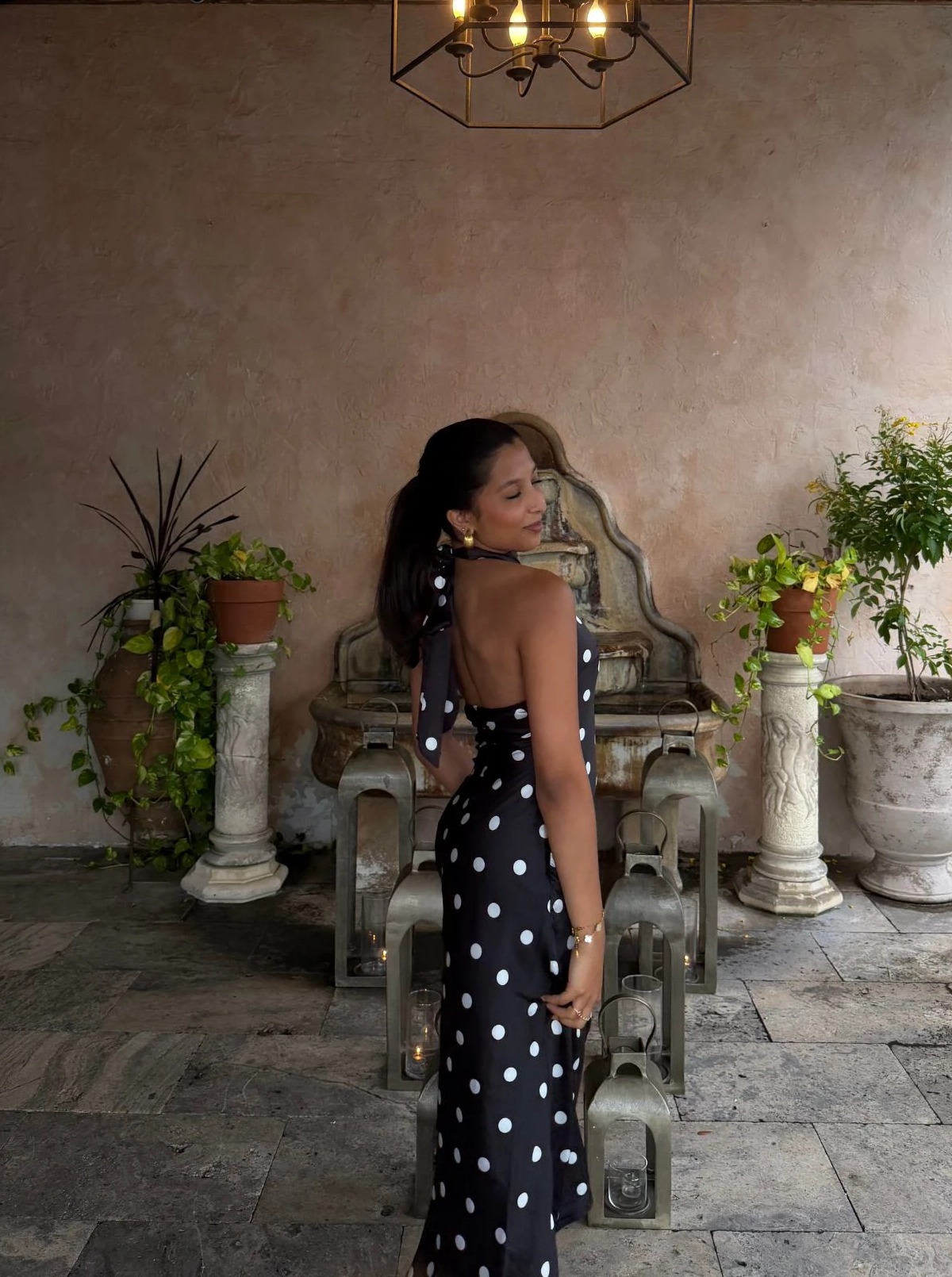 Diya Kodgire standing in a sunlit courtyard in a black and white polka-dot dress, looking back over her shoulder and smiling, with a stone fountain, potted plants, and a hanging chandelier behind her.