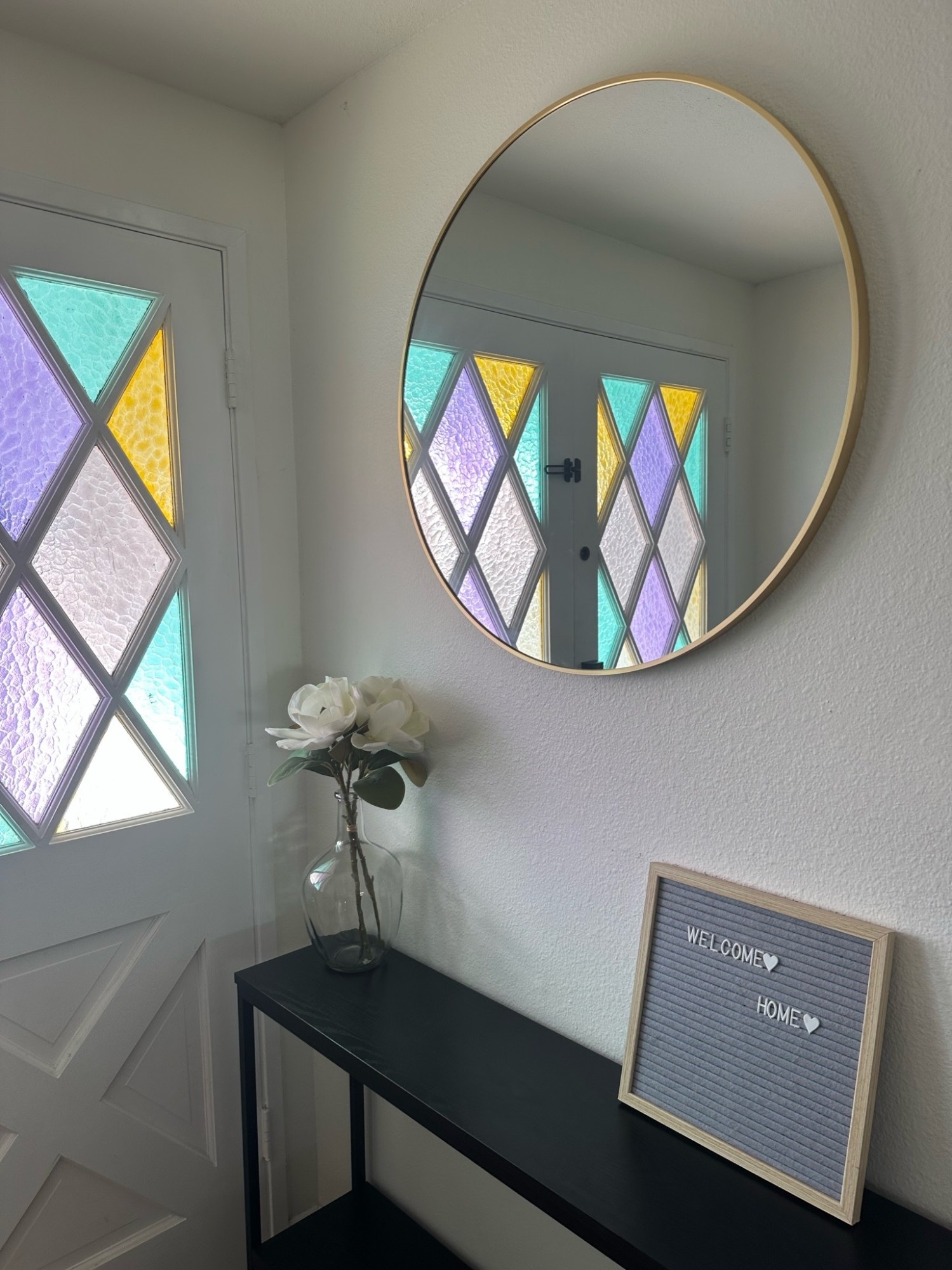 A round gold-framed mirror mounted above a black console table, reflecting a front door with colorful stained-glass diamond panes.