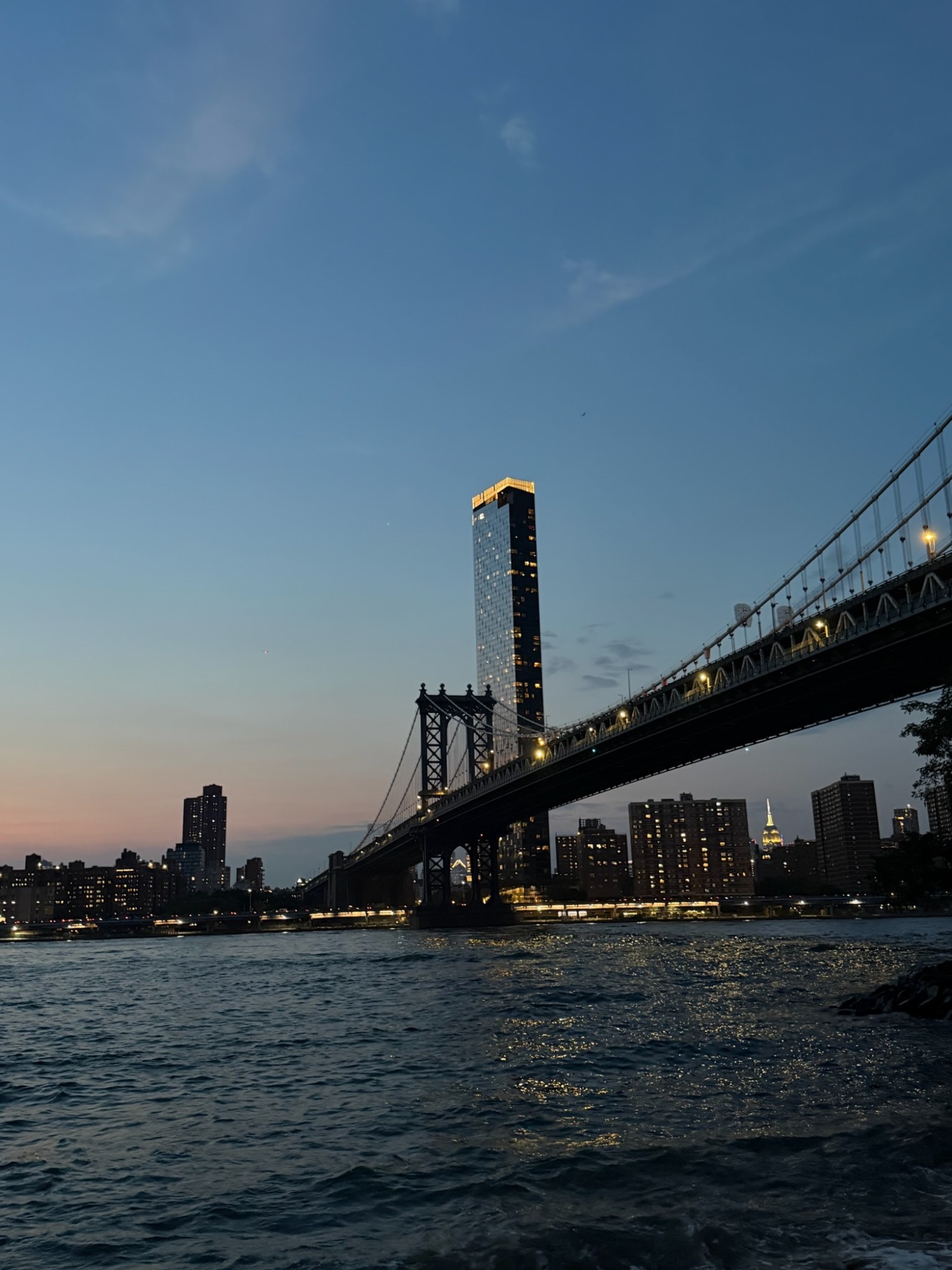 A suspension bridge's stone tower rising over a river at dusk, with a lit high-rise building and a city skyline silhouetted against a pink-to-blue sky.