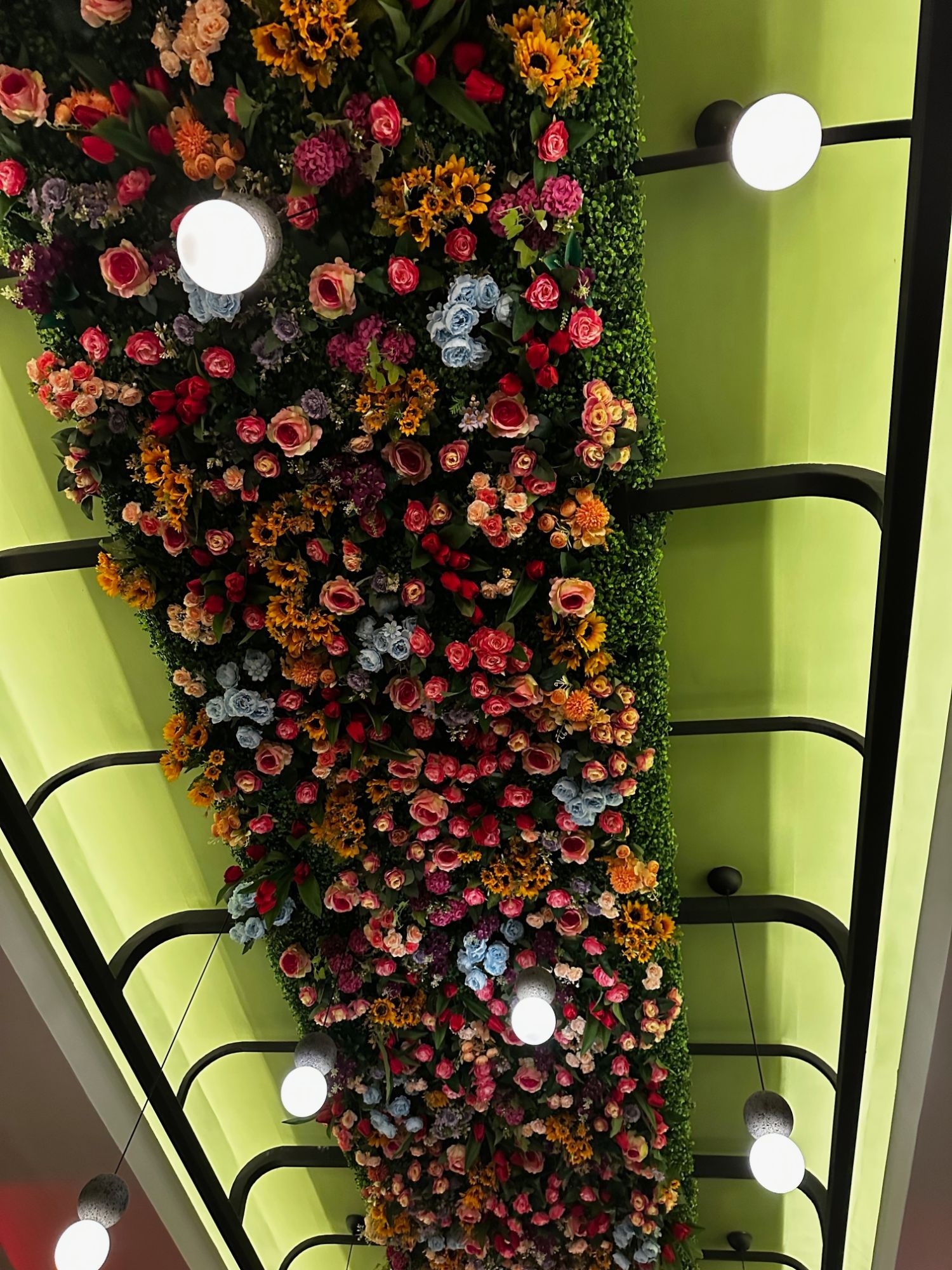 An indoor ceiling and wall densely covered in an array of pink, red, orange, and blue artificial flowers, lit by round pendant lights, with a staircase railing visible below.