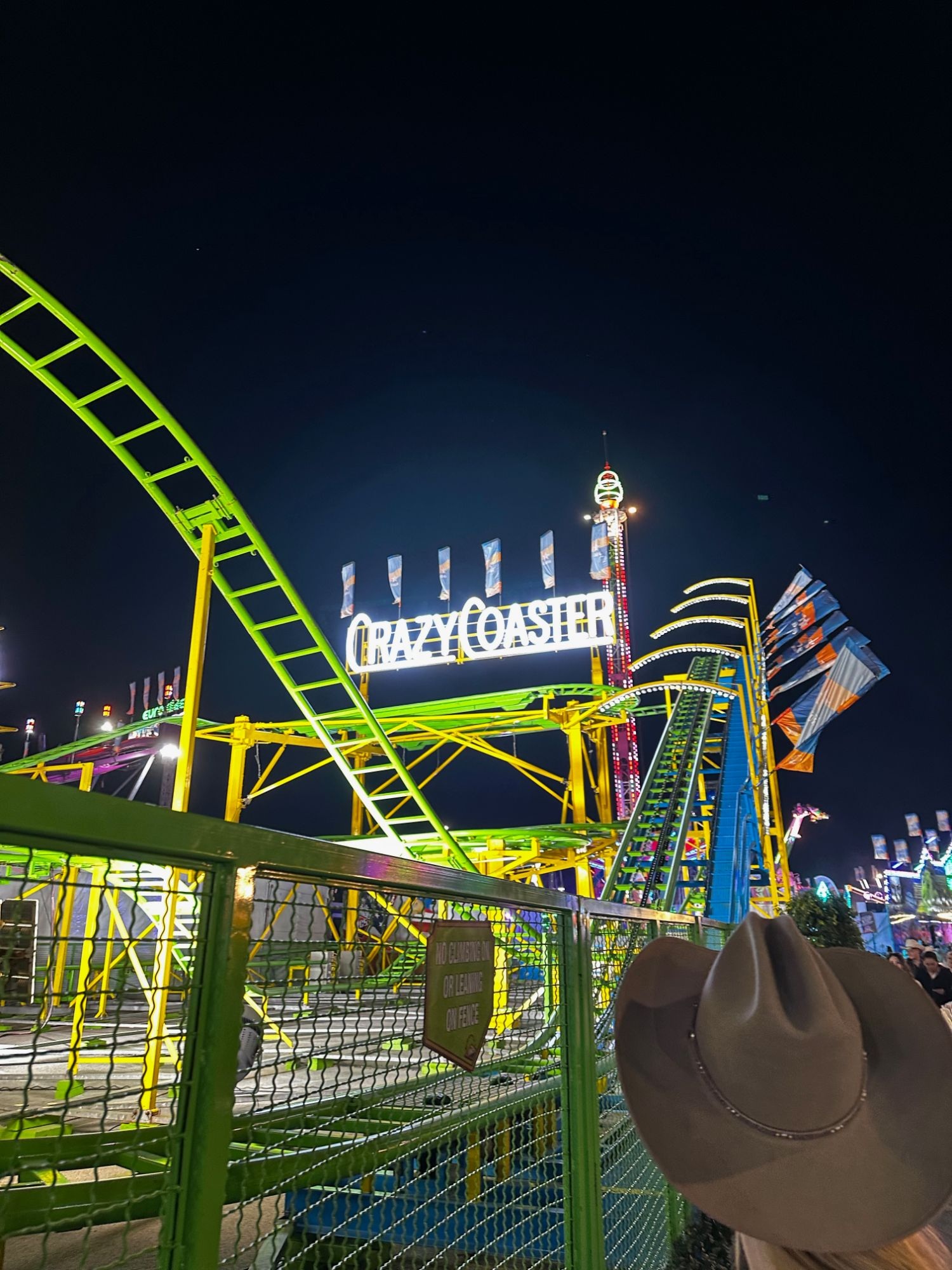 A green roller coaster track lit at night beside a lit Crazy Coaster sign, with a cowboy hat partially blocking the foreground.