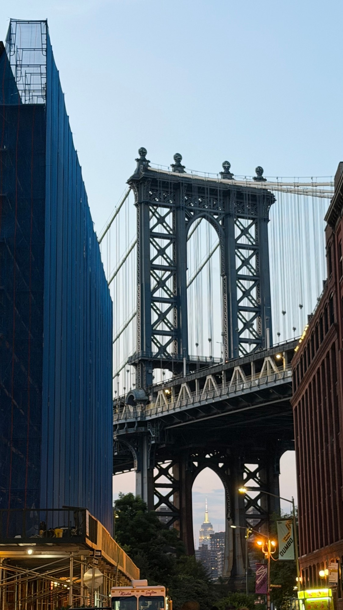 A view of the Manhattan Bridge's stone tower and steel truss roadway from a DUMBO street between two buildings, with the Empire State Building visible through the bridge's arch.