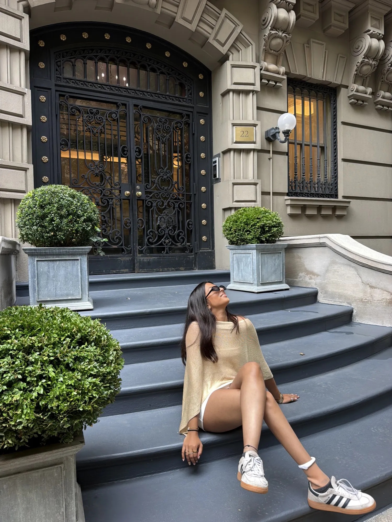 A woman sitting on the stone steps of an ornate townhouse entrance with a wrought-iron door, looking up, flanked by potted boxwood topiaries.