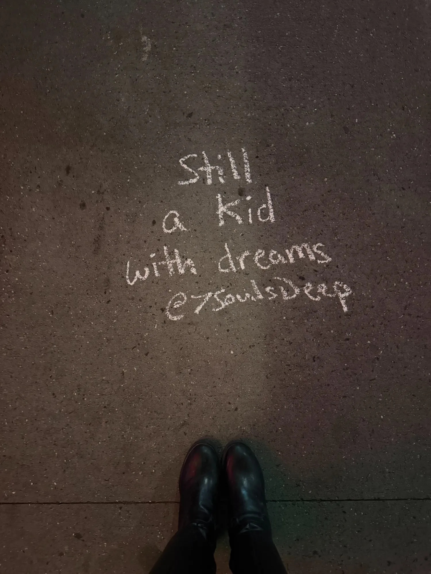 Handwritten chalk text on dark pavement reading Still a kid with dreams at SoulsDeep, with a pair of shoes visible at the bottom of the frame.