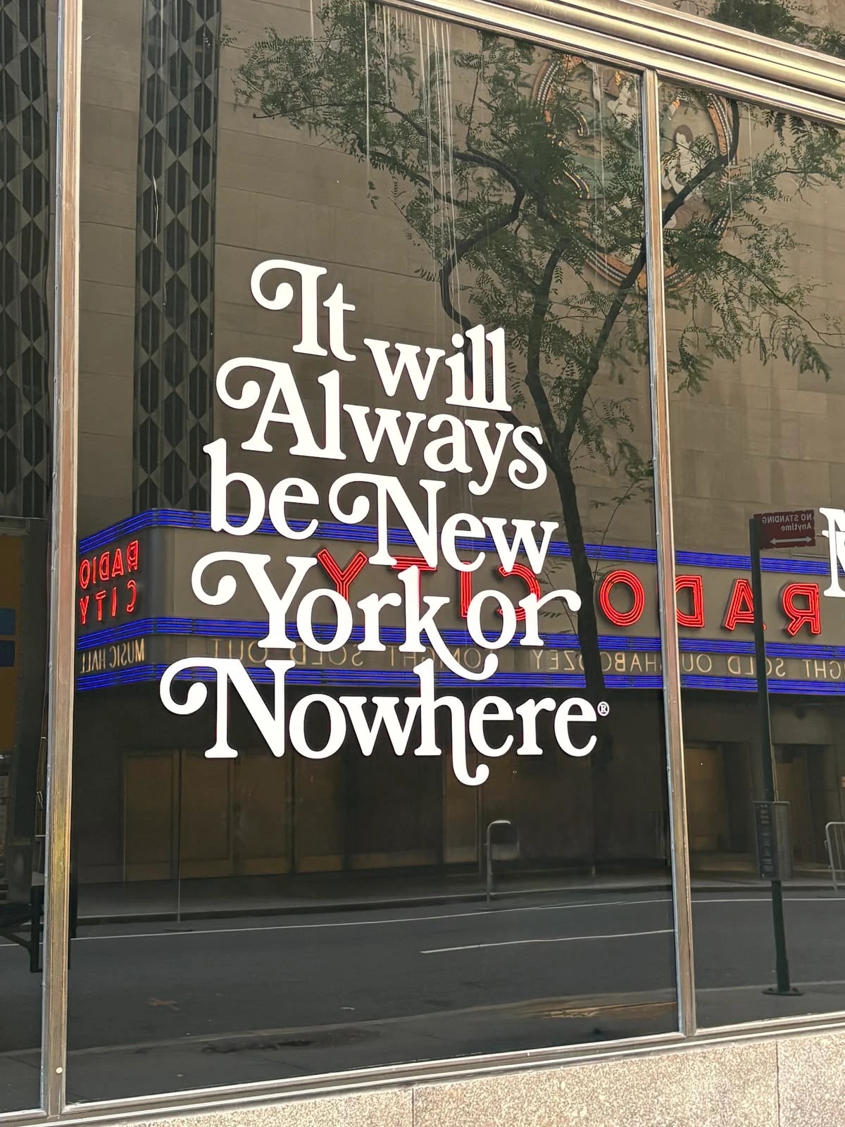 A storefront window with white and red script text reading It will Always be New York or Nowhere, reflecting a theater marquee reading Radio City Music Hall across the street.