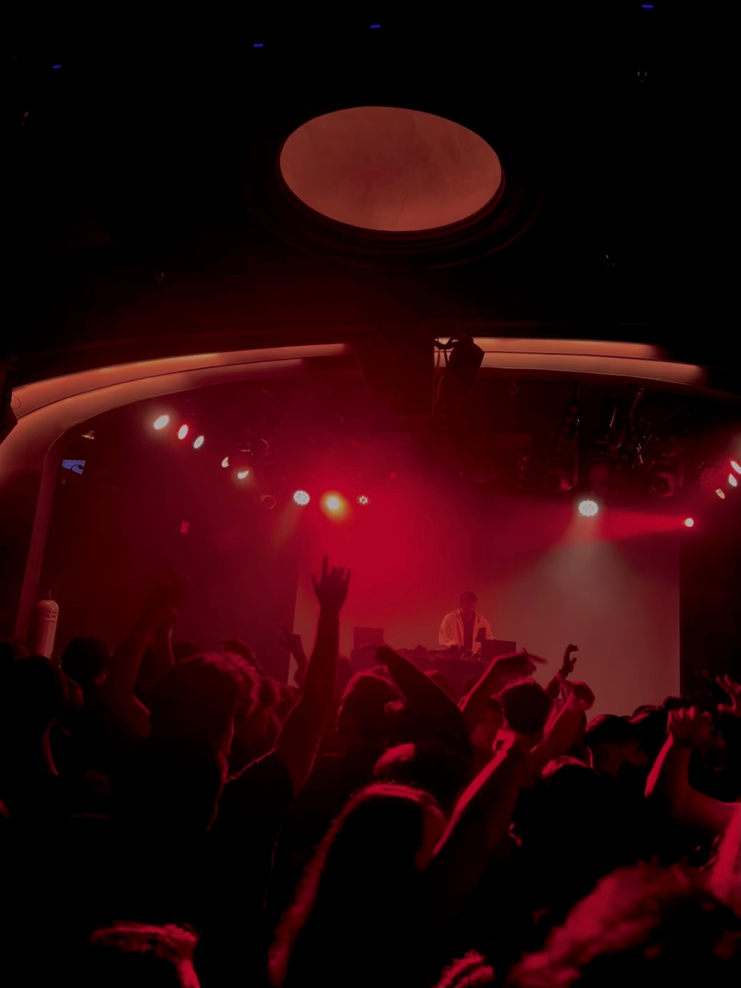 A concert crowd with raised hands silhouetted in red stage lighting, facing a DJ performing under a round lighting rig.