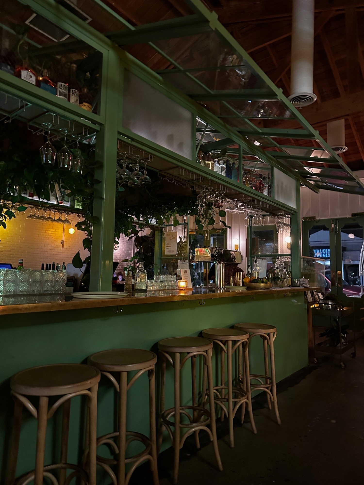 An empty green-painted bar counter with wooden stools, hanging wine glasses, and potted plants, lit warmly at night.