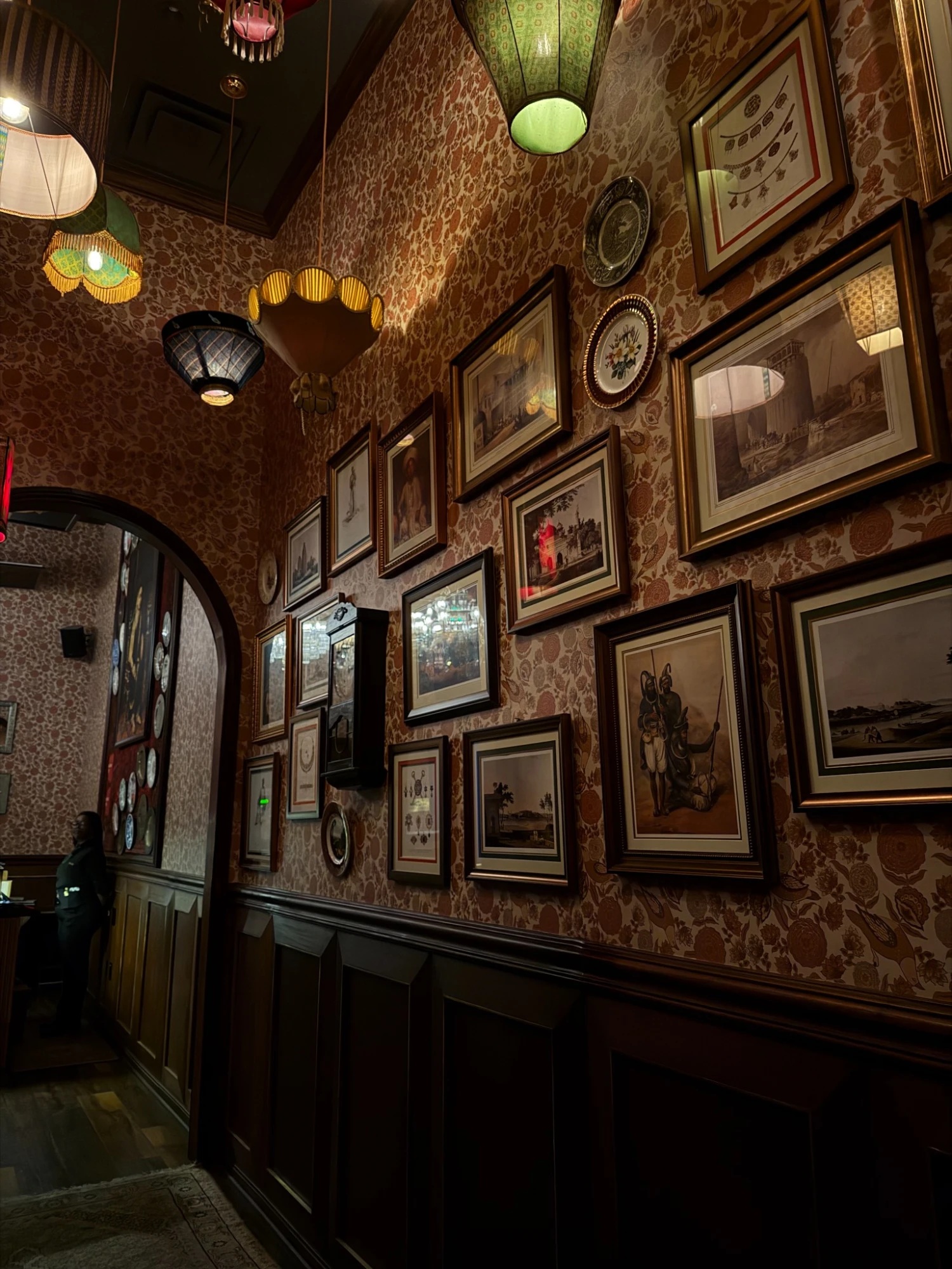 A wall densely hung with ornately framed vintage prints and decorative plates, under patterned wallpaper and colorful hanging lampshades.