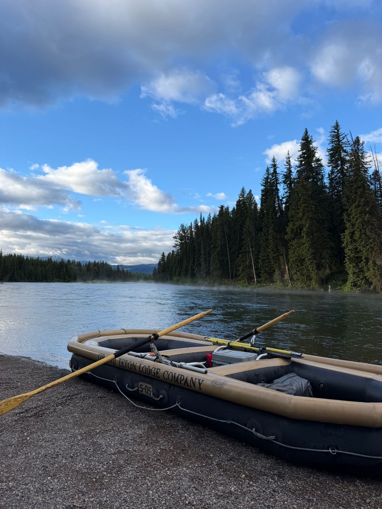 An inflatable river raft with oars resting on a gravel riverbank, facing a calm river lined with pine trees under a partly cloudy sky.