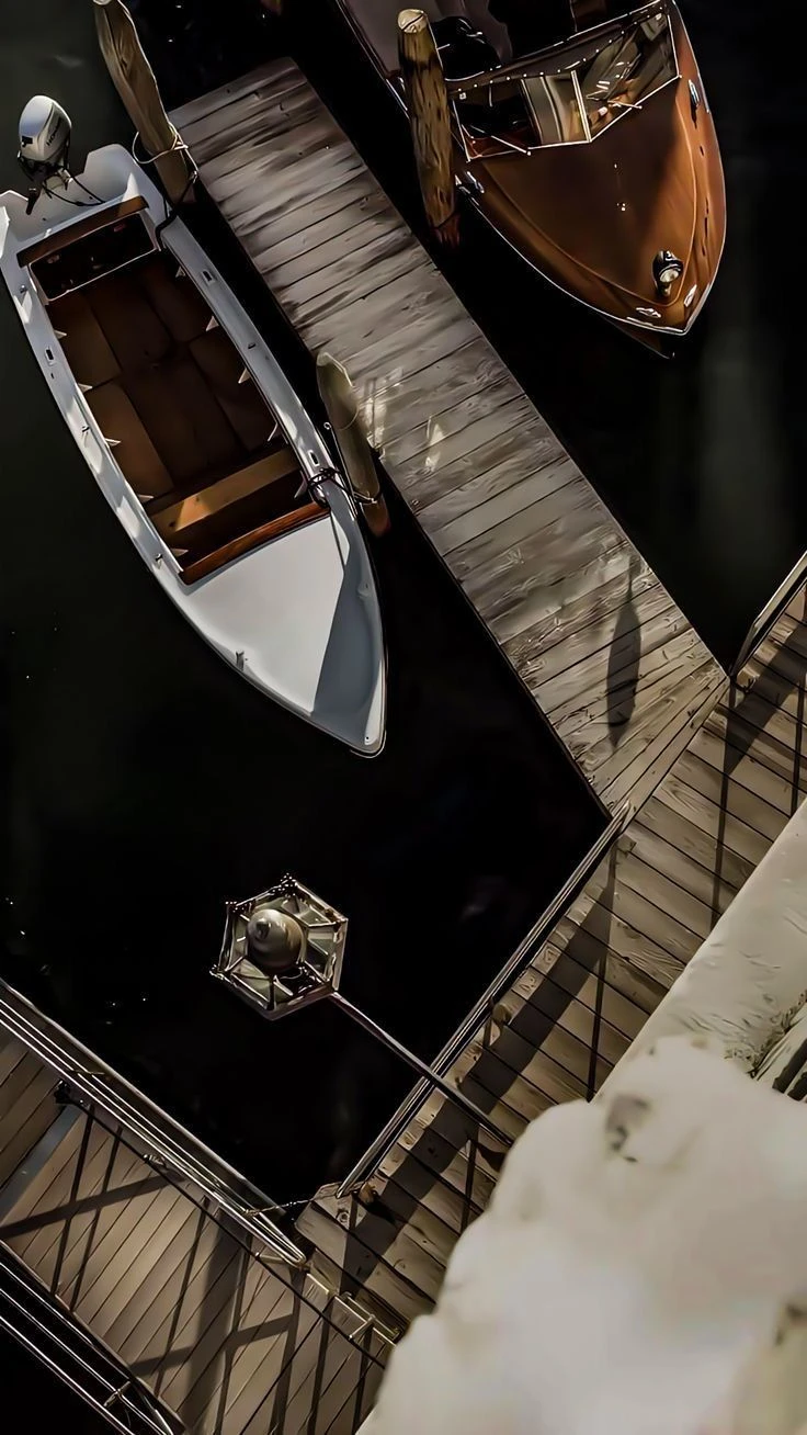 Overhead view of two wooden motorboats moored at a sunlit dock, one white-hulled, one varnished mahogany, with a lantern post and railing in shadow below.